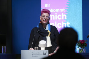 Auf dem Bild ist die ungehaltene Rednerin Sandra Kossendey zu sehen, sie schaut ernst ins Publikum, ihre kurzen Haare sind pink gefärbt.