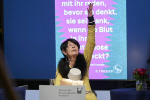 Auf dem Bild ist die Mitte 80jährige ungehaltene Rednerin Eva Schulz-Jander in einem gelben Oberteil zu sehen, sie reckt während ihrer Rede eine Hand in die Höhe.